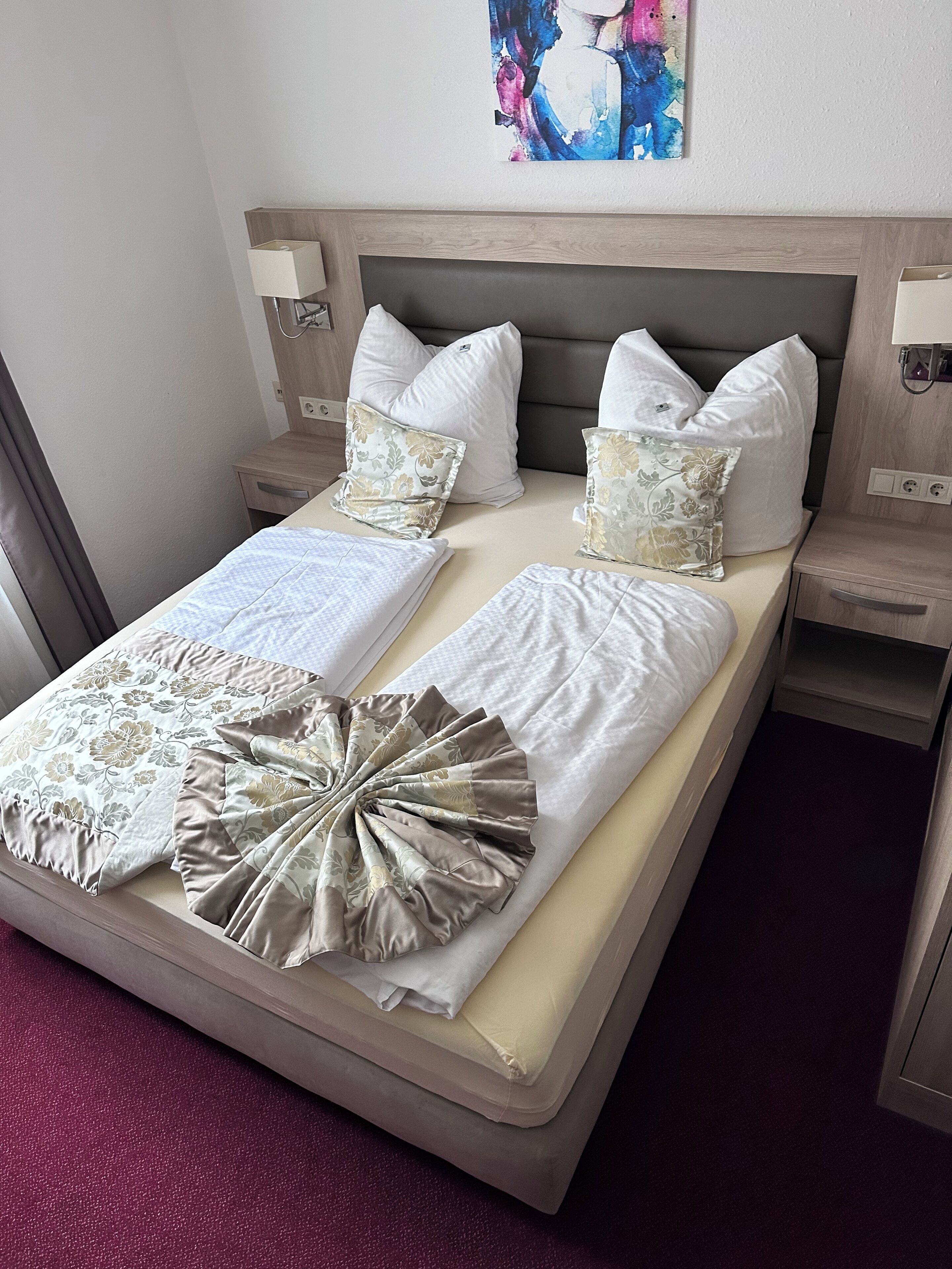 Ein ordentlich bezogenes Bett mit weißen Leinen und dekorativen Kissen, ein Nachttisch mit einer Lampe und ein Gemälde über dem Bett.