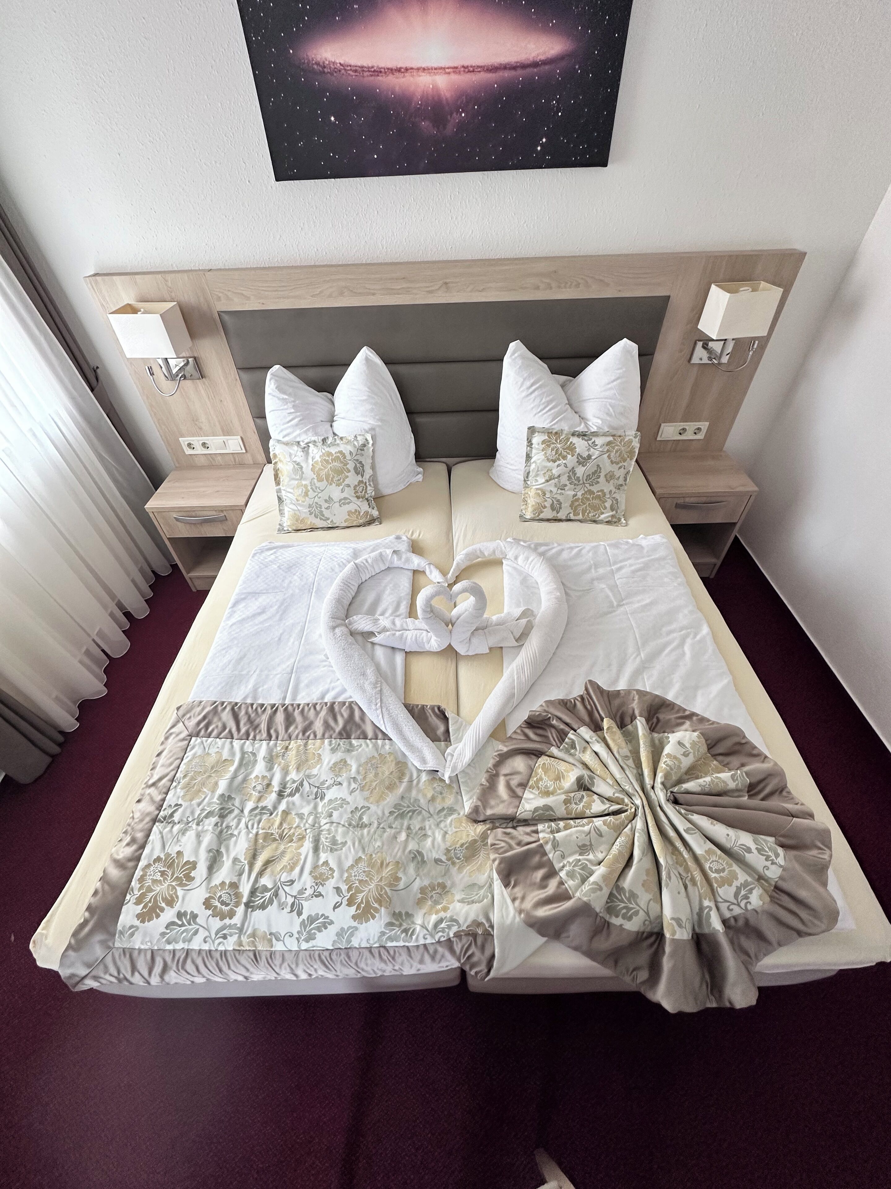 Een netjes opgemaakt bed met beddengoed met bloemenprint, een handdoekje gevouwen in de vorm van een hart, en een wanddecoratie met een sterrenstelsel thema erboven.