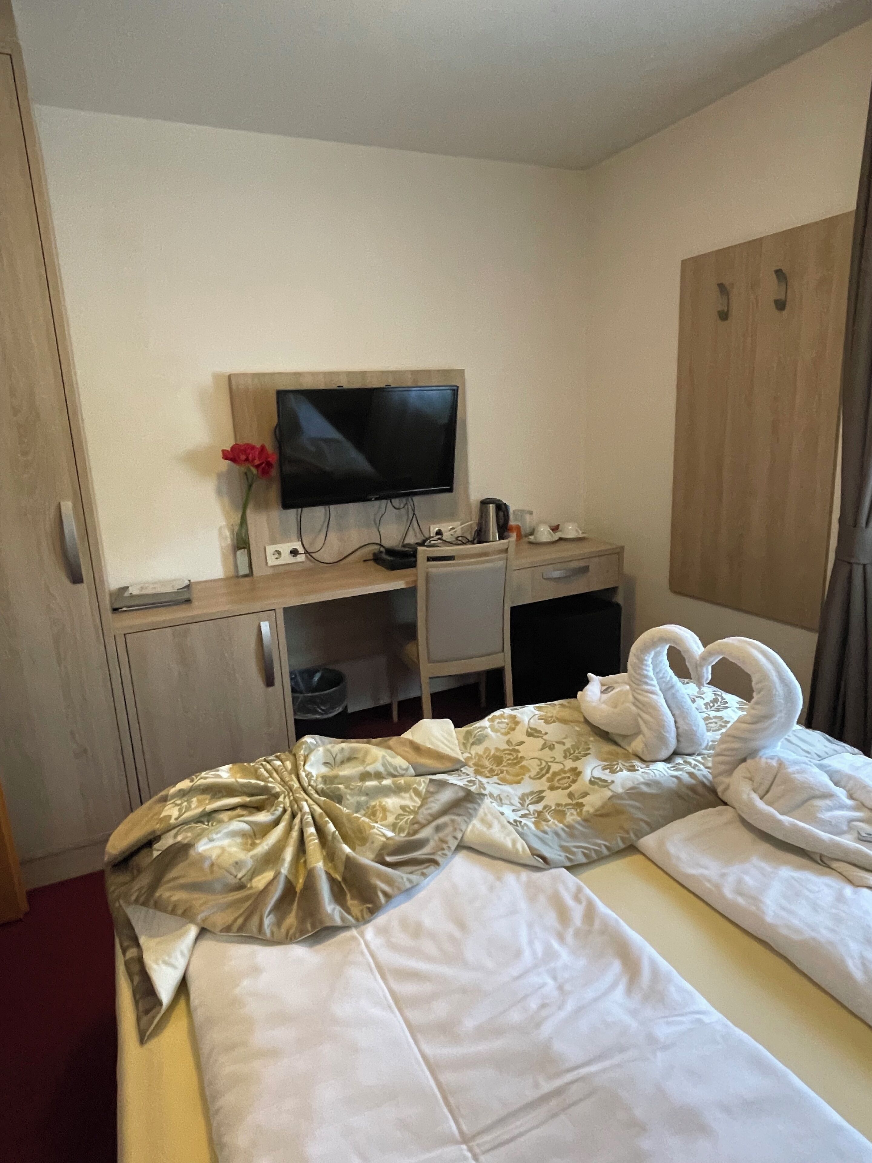 Ein Hotelzimmer mit einem Bett, einem Schreibtisch mit Fernseher, einem Kleiderschrank und einer roten Rose auf dem Schreibtisch.