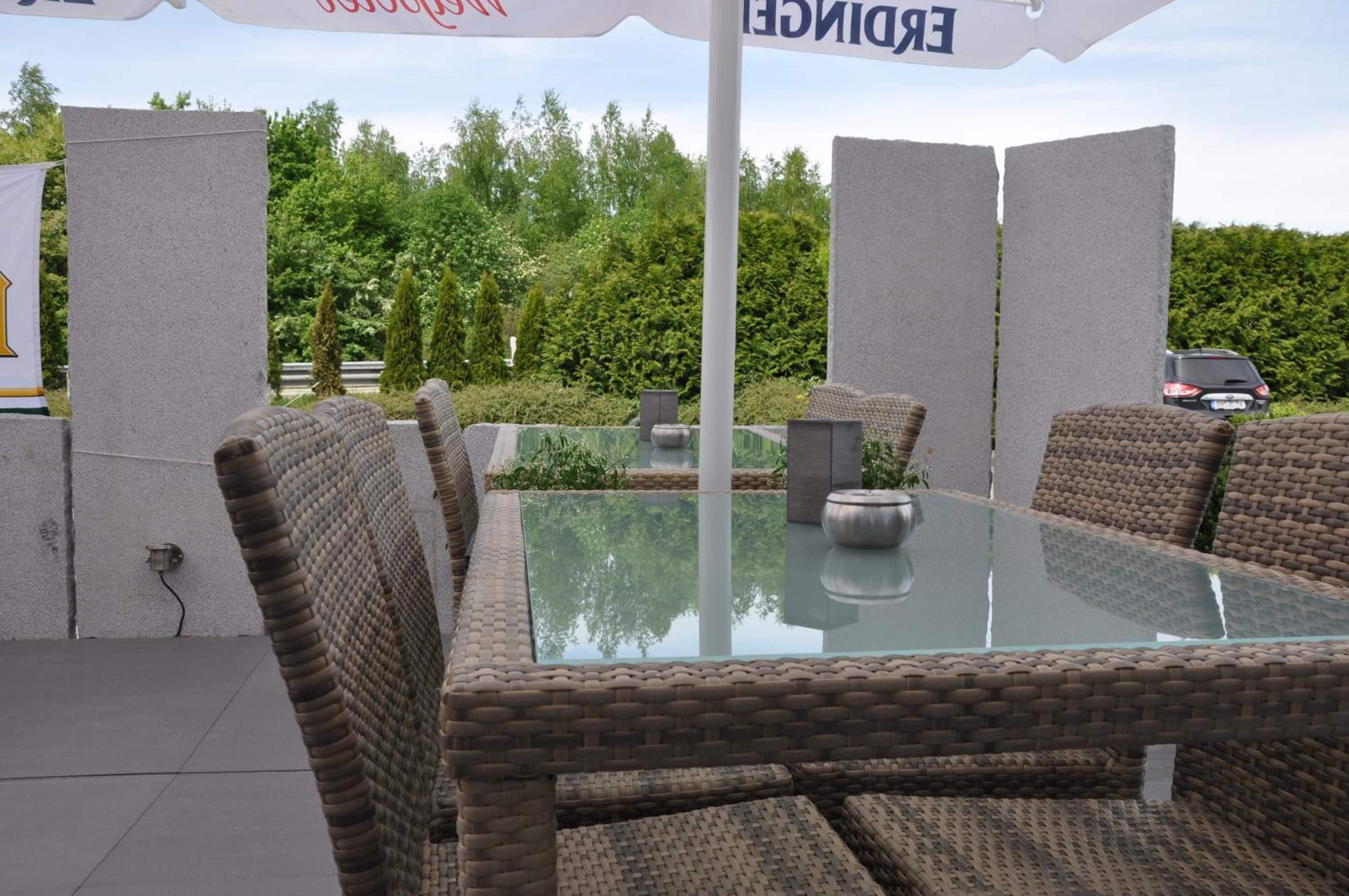 An outdoor seating area with wicker chairs and a glass table, surrounded by a hedge and a parked car.