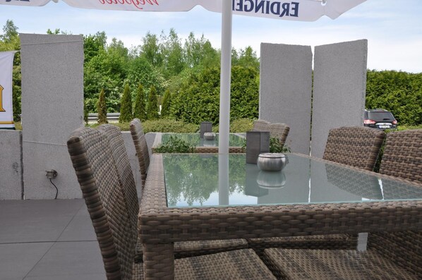 An outdoor seating area with wicker chairs and a glass table, surrounded by a hedge and a parked car.