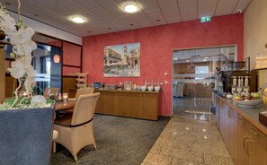 Un hall d’hôtel comprenant un comptoir d’accueil, un espace repas avec des chaises en osier, un grand tableau accroché au mur et une cuisine visible à travers une cloison en verre.