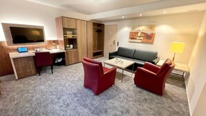 Habitación de hotel con televisor de pantalla plana, escritorio con computadora portátil, sofá, dos sillas rojas, mesa de centro y un cuadro en la pared.