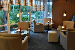 A hotel room with a seating area featuring armchairs and a small round table, large windows with a view of greenery, and a carpeted floor.