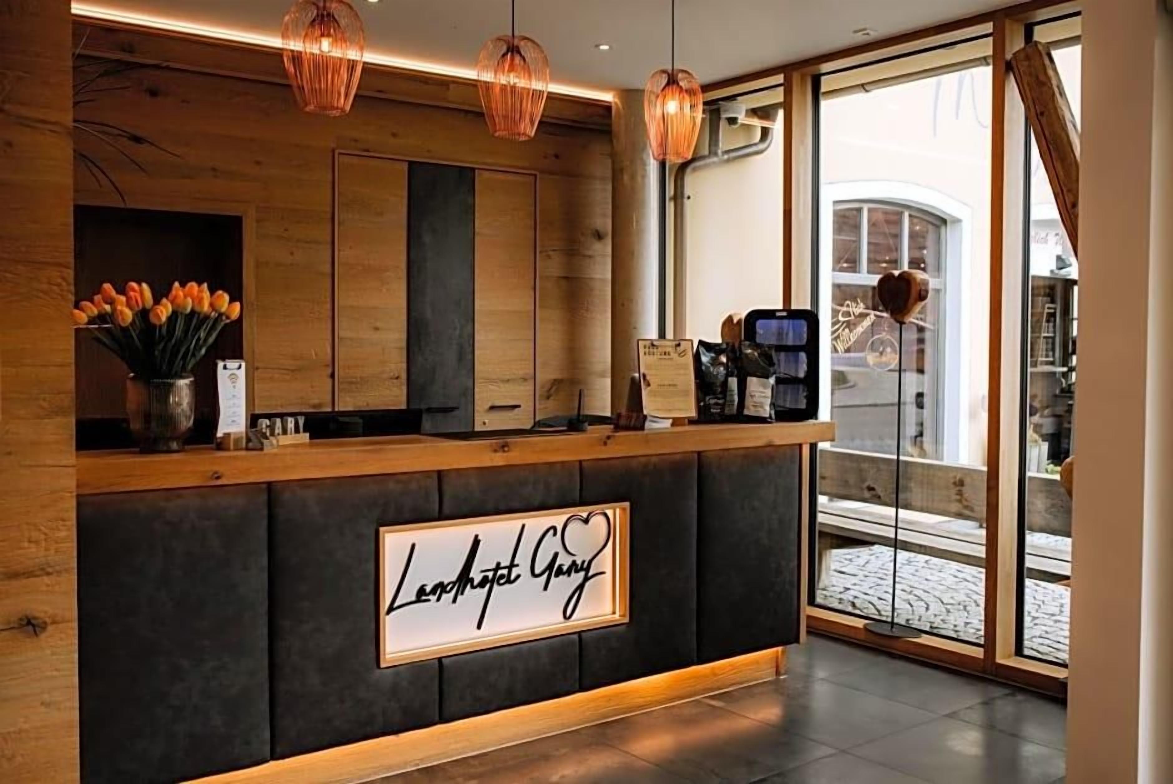 Un comptoir d’accueil avec une pancarte indiquant « Lennapet Gary » et un symbole en forme de cœur. Au-dessus du comptoir, on aperçoit des panneaux en bois, et à gauche, un vase rempli de fleurs oranges.