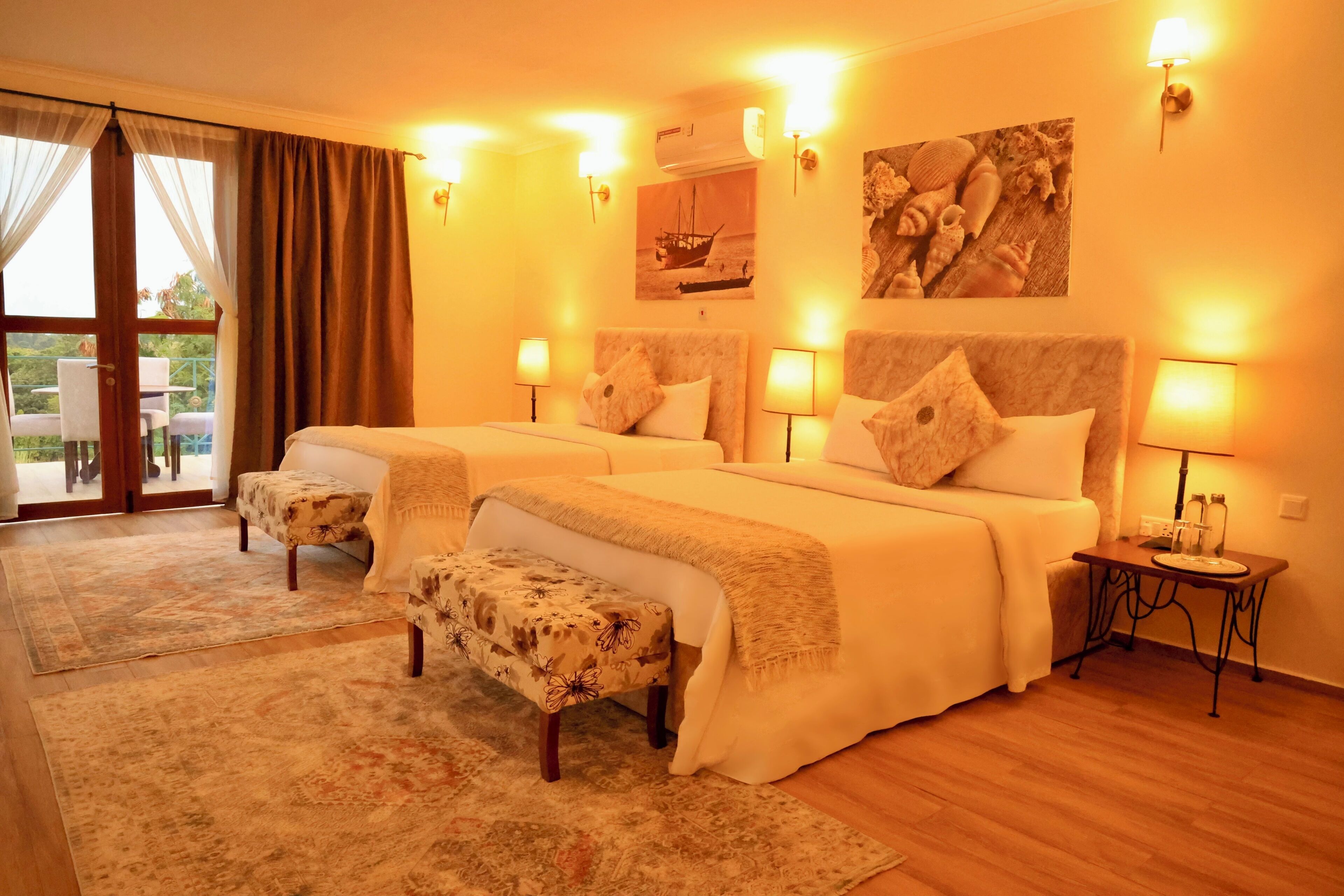 A hotel room with two beds, a bench, a small table, and a view through a glass door.