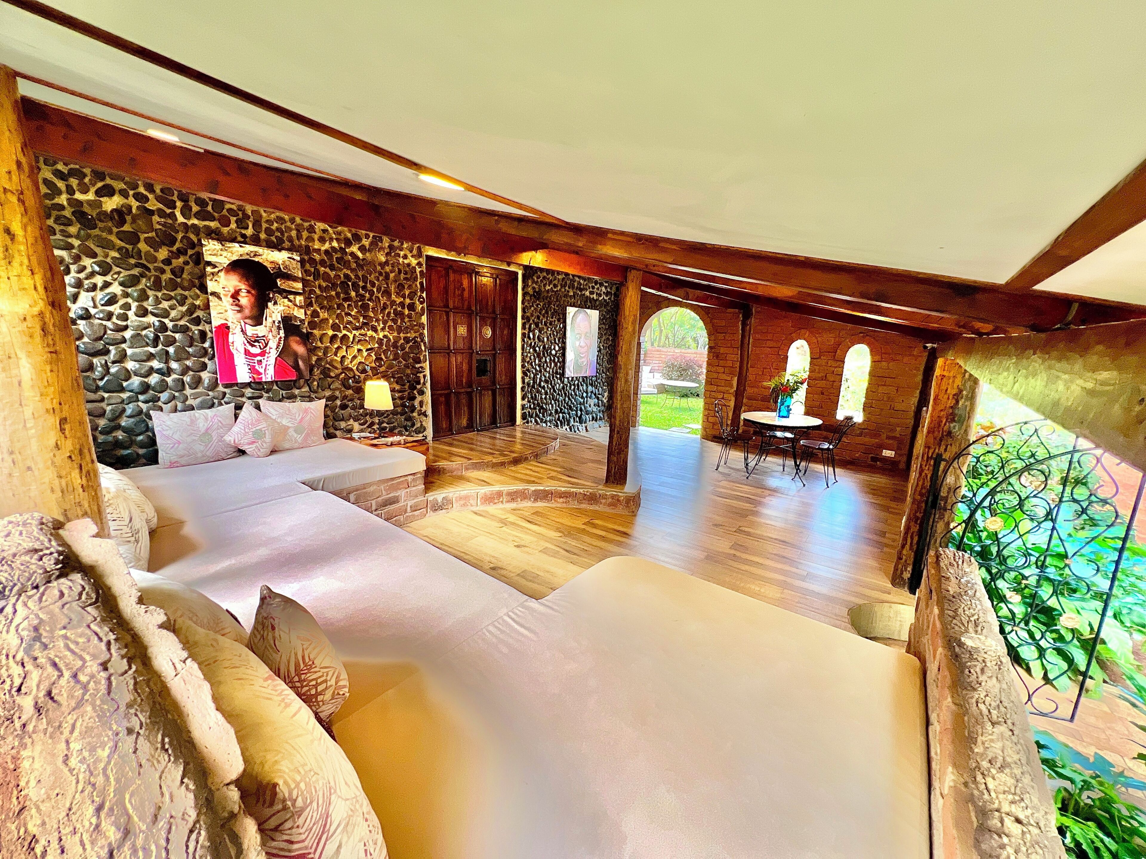 A spacious room with a stone wall, wooden beams, and a large bed with multiple pillows. There is a table with chairs and a view of the outside.