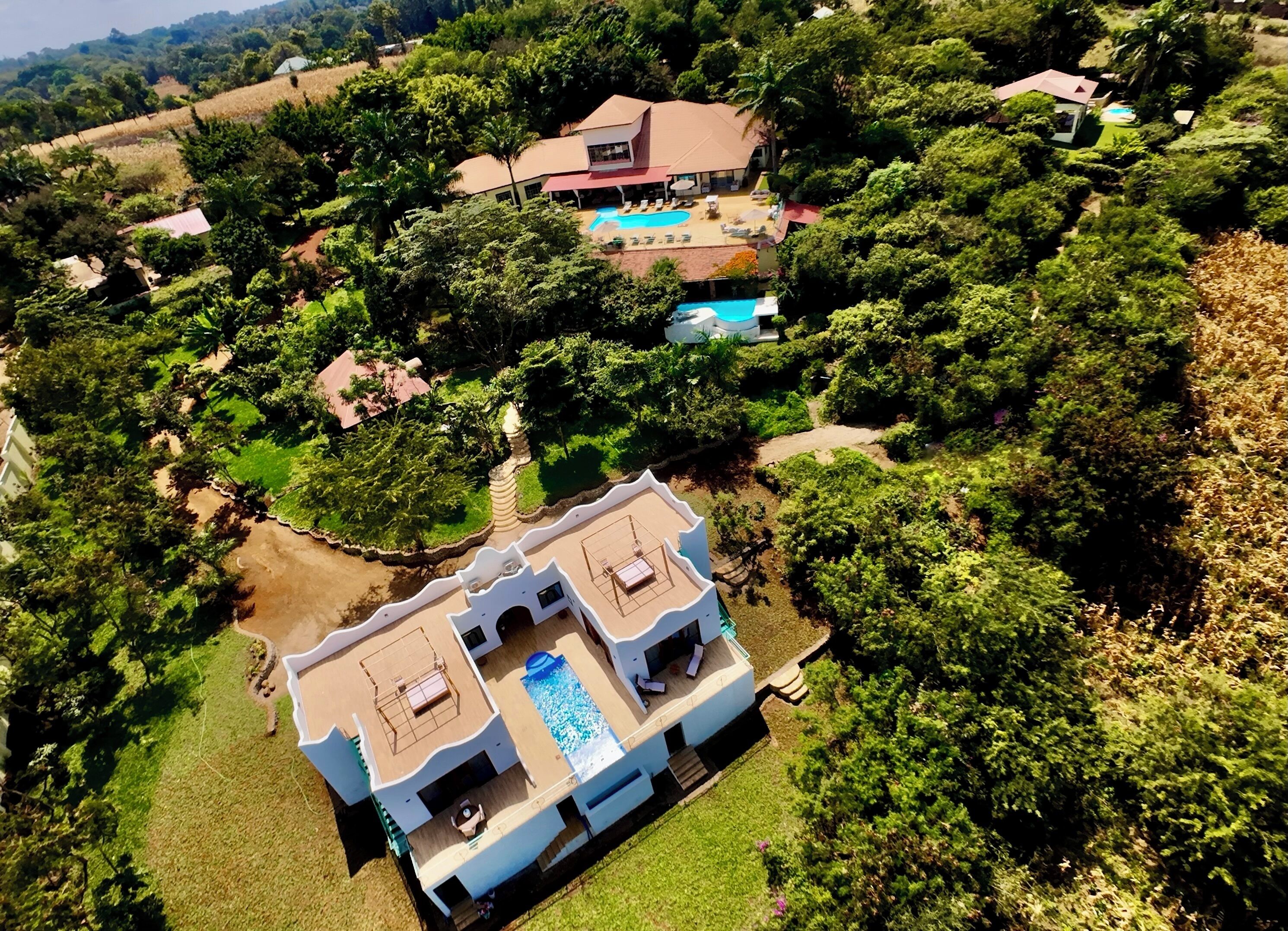 An aerial view of a large, modern house with a swimming pool, surrounded by lush greenery and a hilly landscape.