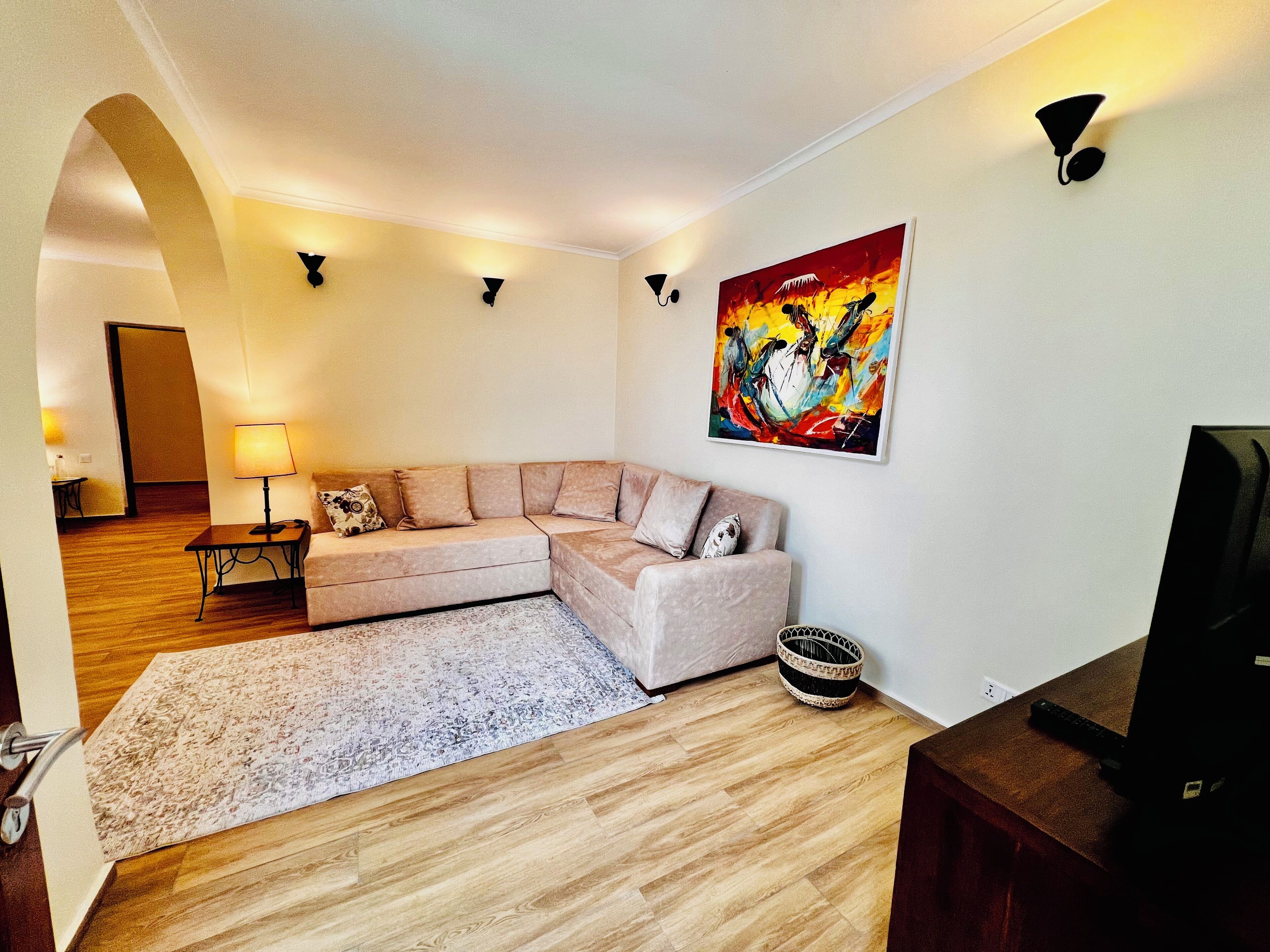 A modern living room with a beige sectional sofa, a flat-screen TV, a colorful abstract painting, and a small side table with a lamp.