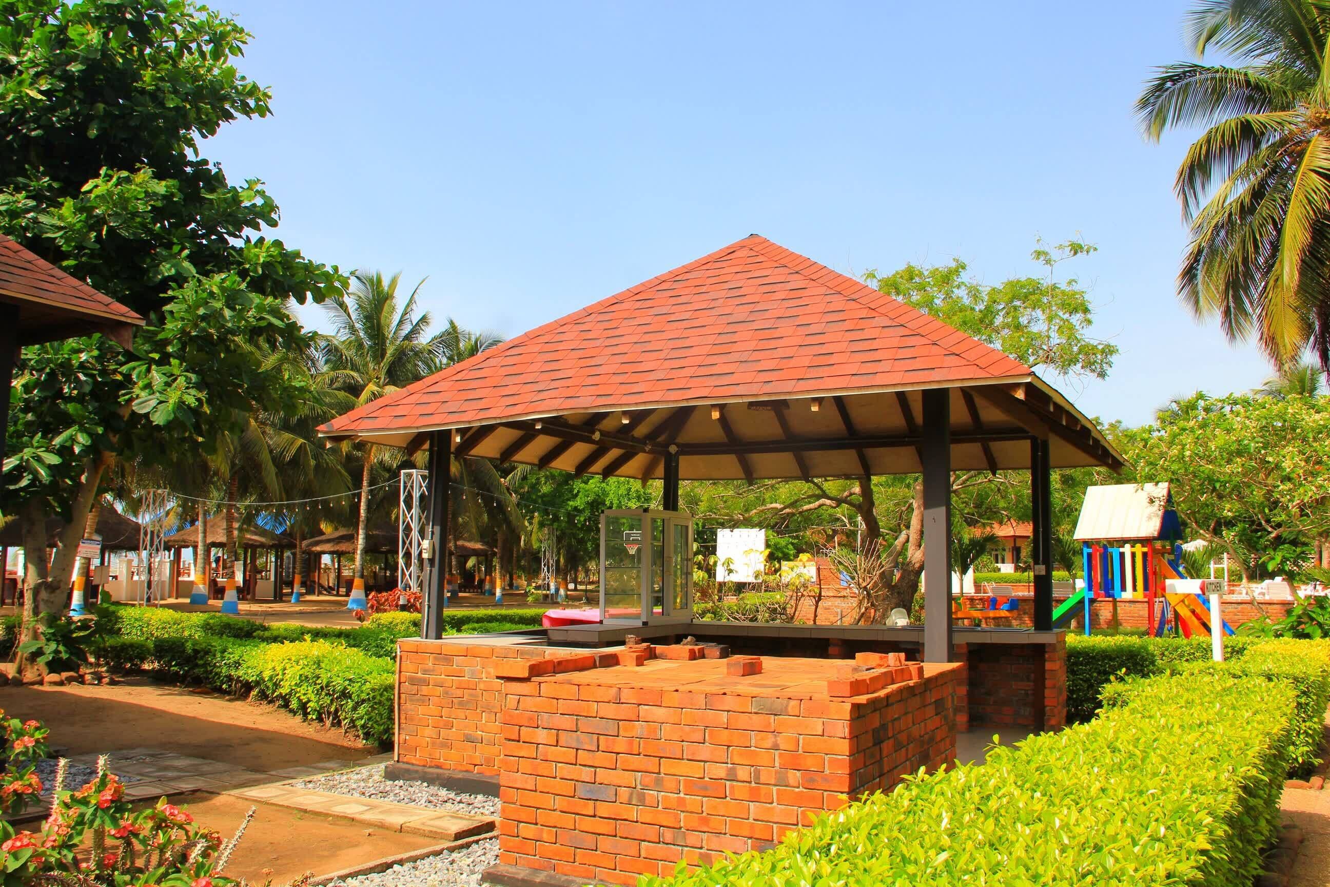 A brick pavilion with a tiled roof, surrounded by well-maintained hedges and palm trees.