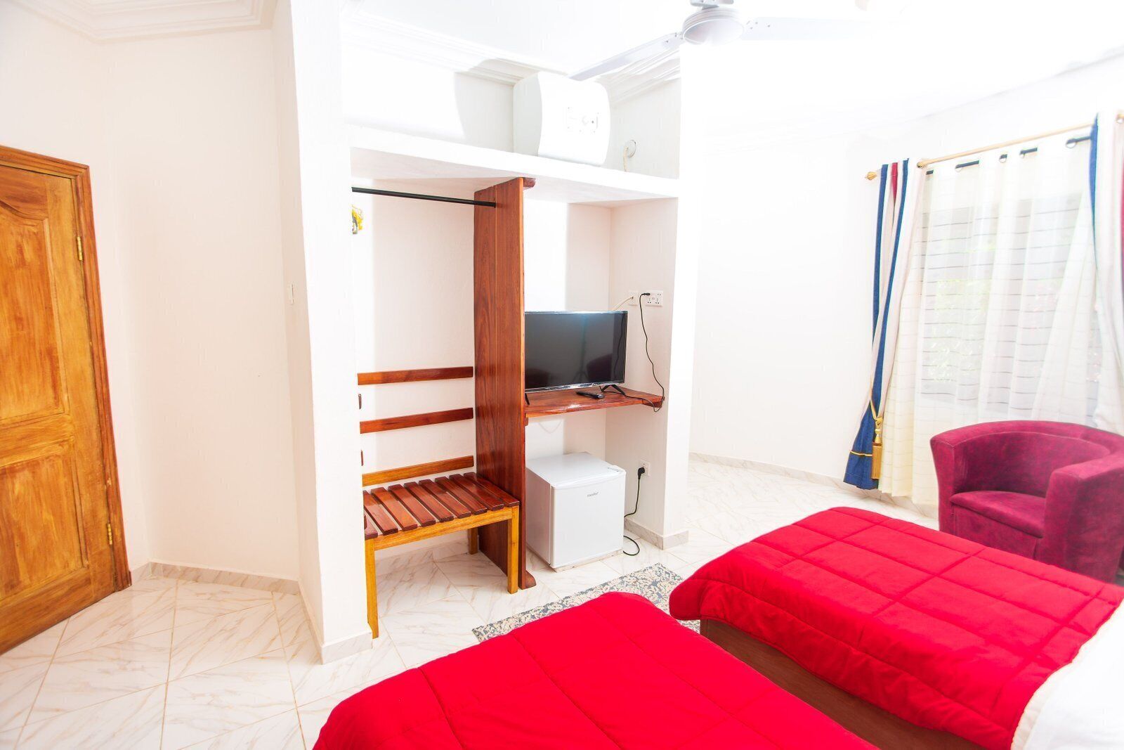 A room with a bed, a desk with a computer, a wardrobe, a red chair, and a window with curtains.