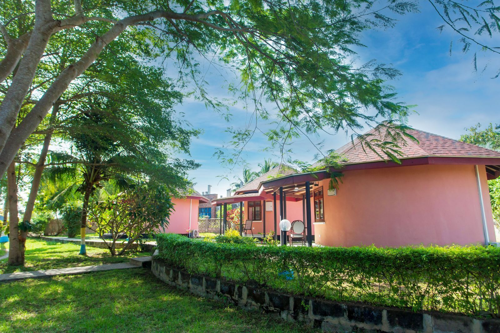 A pink building with a covered patio area, surrounded by lush greenery and a stone wall.