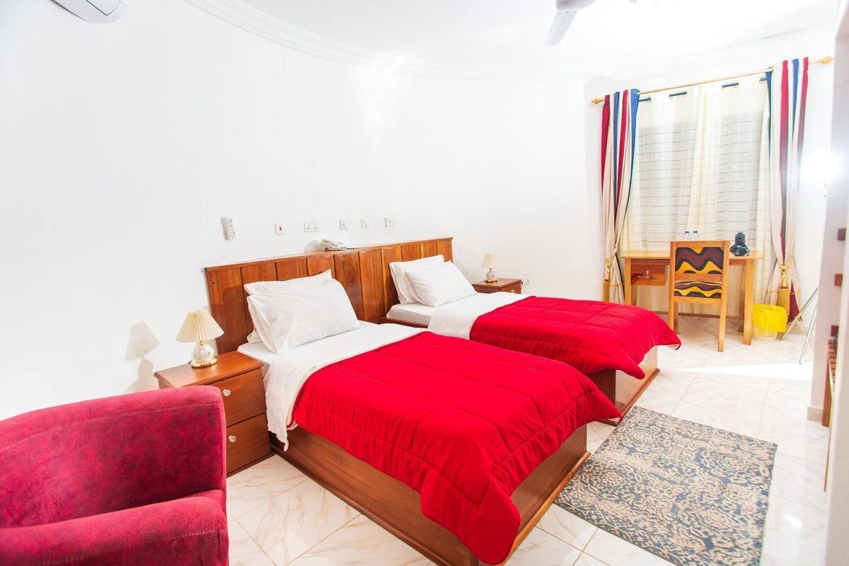 A double bed with red bedding, a wooden nightstand with a lamp, a red chair, and a small desk with a chair.