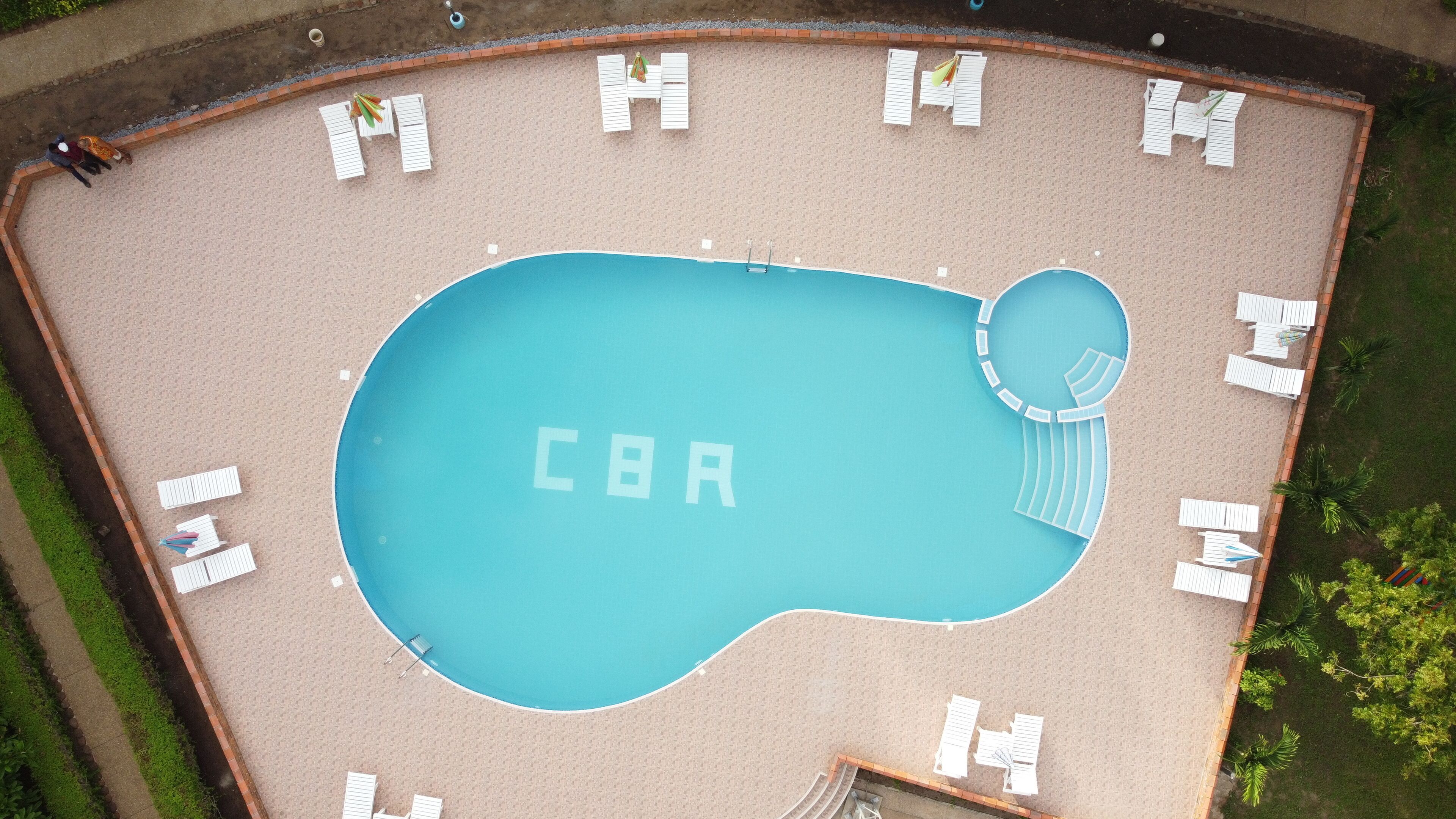 Eine Vogelperspektive auf einen Swimmingpool, in dessen Mitte 'CBA' steht, umgeben von Liegestühlen und einem Mauerwerk-Rand.