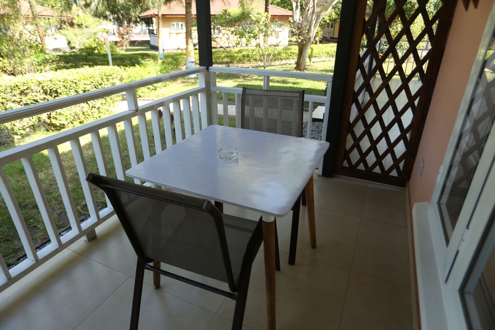 A balcony with a white table and two chairs, a view of a garden, and a lattice door.