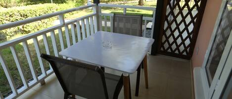 A balcony with a white table and two chairs, a view of a garden, and a lattice door.