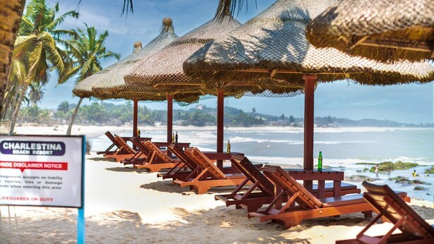 A beach with wooden lounge chairs and thatched umbrellas, a sign for Charleston Beach Resort, and lifeguards on duty. - Charlestina Beach Resort (Ampenyi)