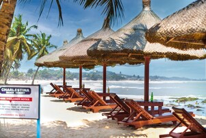 A beach with wooden lounge chairs and thatched umbrellas, a sign for Charleston Beach Resort, and lifeguards on duty.