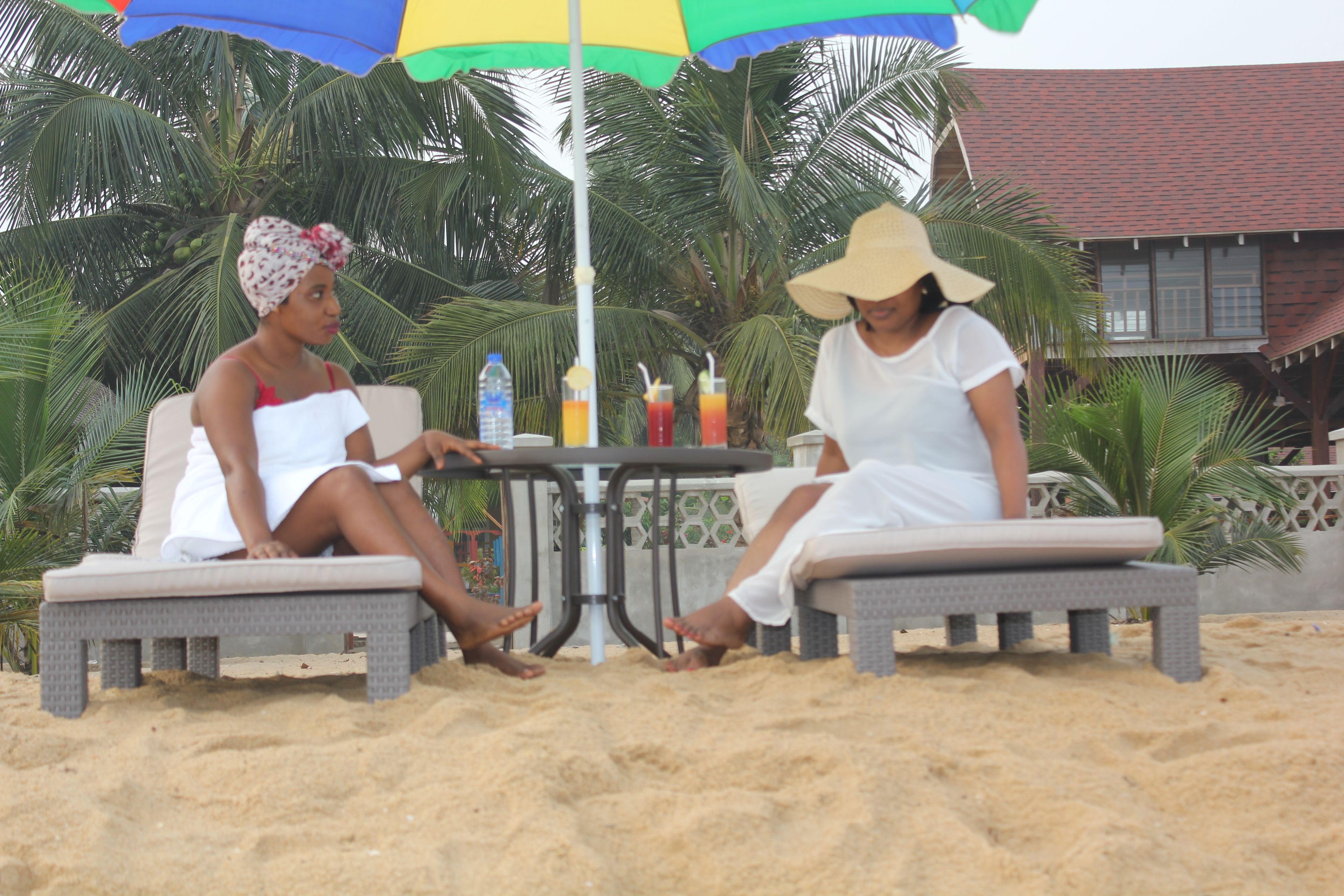 Two people sitting on lounge chairs under a colorful umbrella on a sandy beach with palm trees in the background.