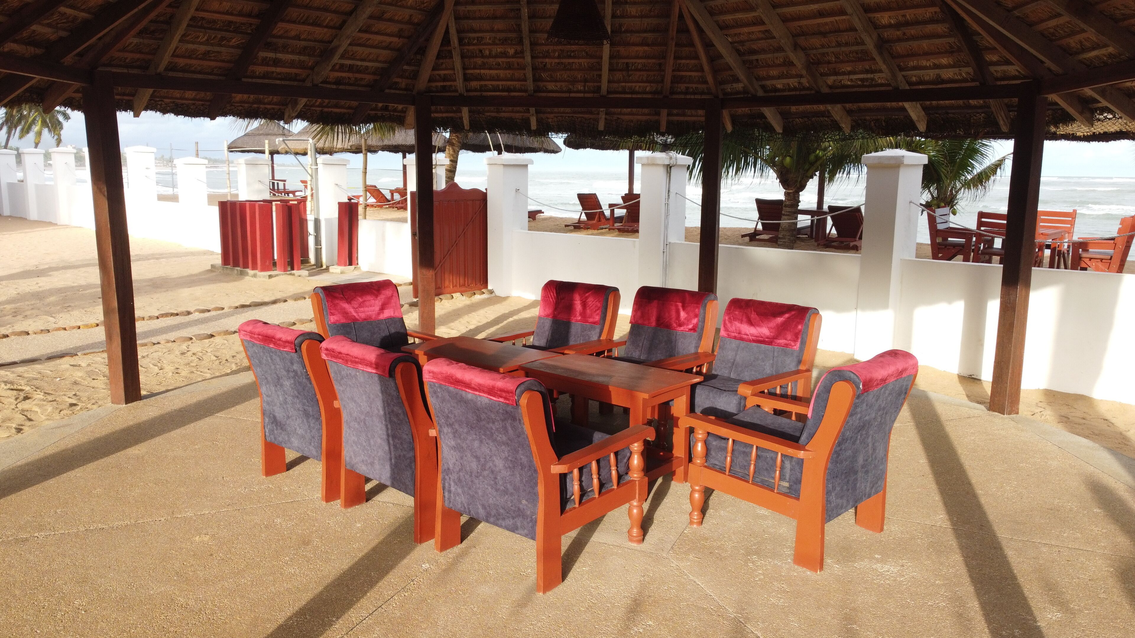 Un área de comedor junto a la playa con una mesa y sillas de madera, techo de paja y vista al océano.
