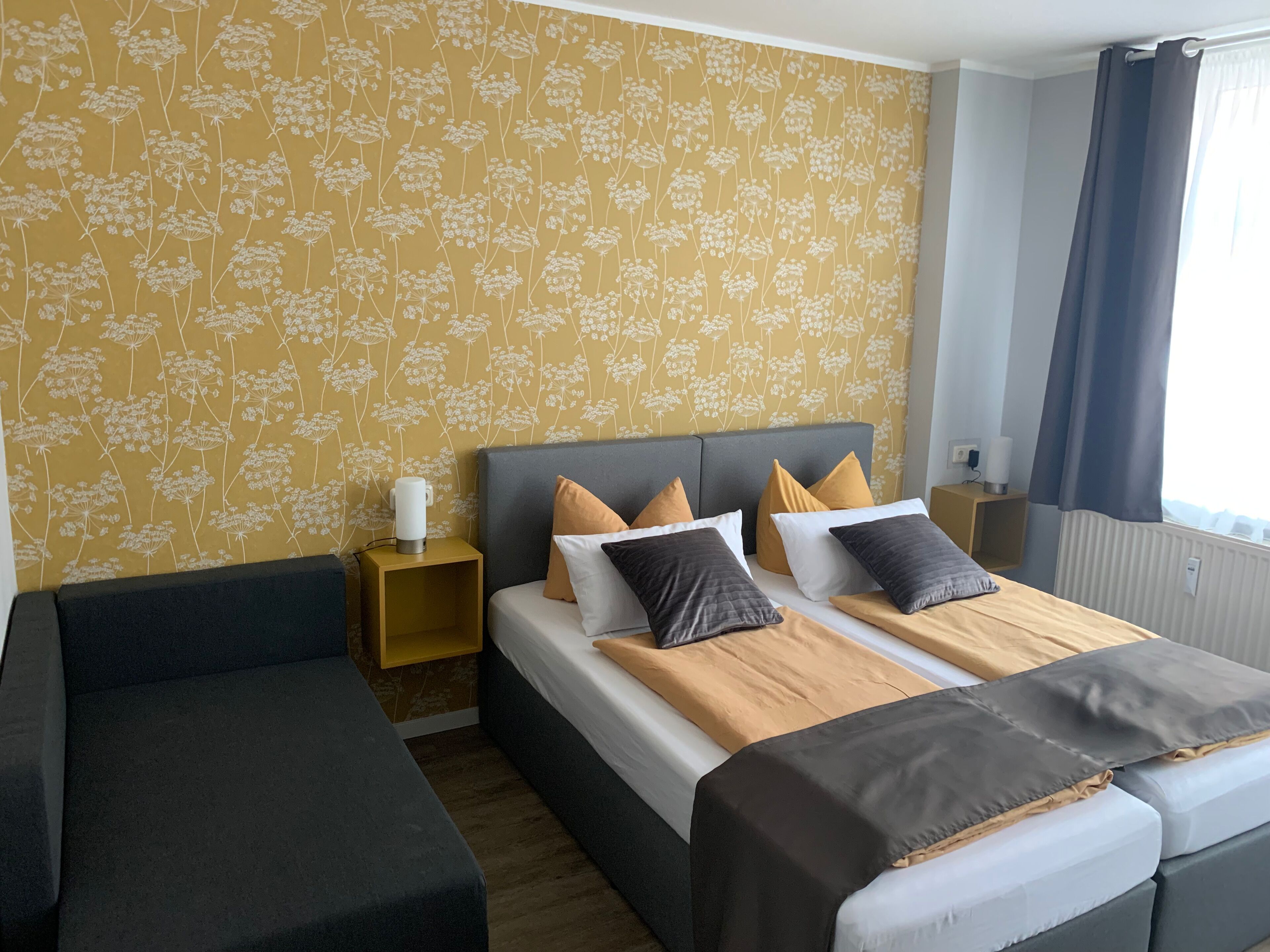 A hotel room with a double bed, two bedside tables, a nightstand, a sofa, and a window with curtains.
