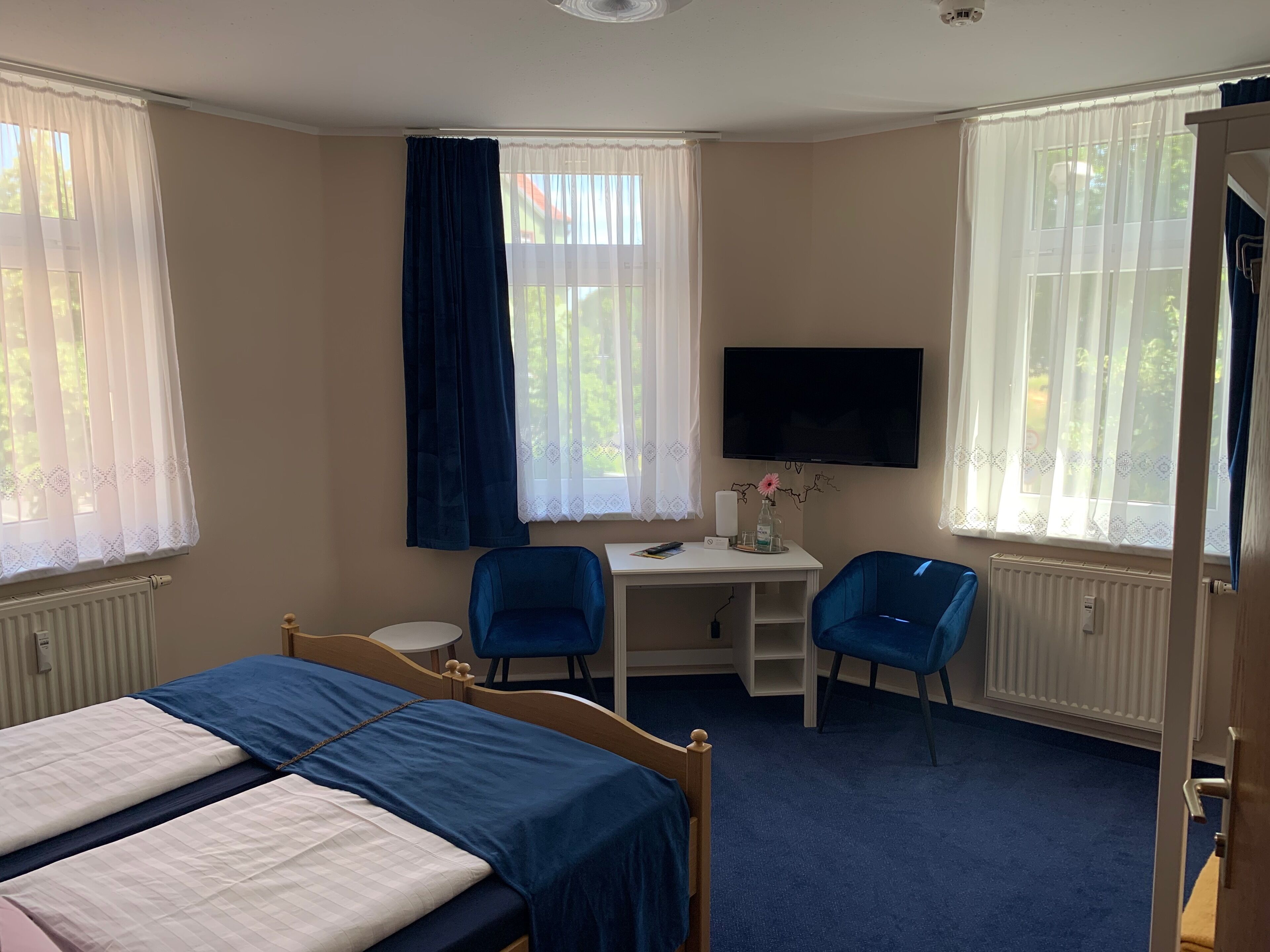 A hotel room with a bed, two chairs, a desk with a TV, and large windows with curtains.