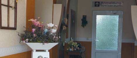 A hallway with a welcome sign, a flower arrangement, and a door labeled "Anmeldung Speiseraum".