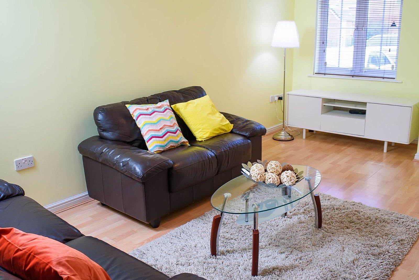Ein Wohnzimmer mit einem dunkelbraunen Ledersofa, einem Glastisch mit Dekoration, einer weißen TV-Konsole und einer Lampe.