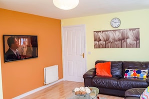 A living room with a television, a door, a radiator, a clock, and a couch with colorful pillows. - Crystal (Manchester)