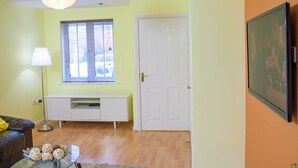 A room with a white door, a window with blinds, a white cabinet, a black sofa, a lamp, and a TV on the wall.