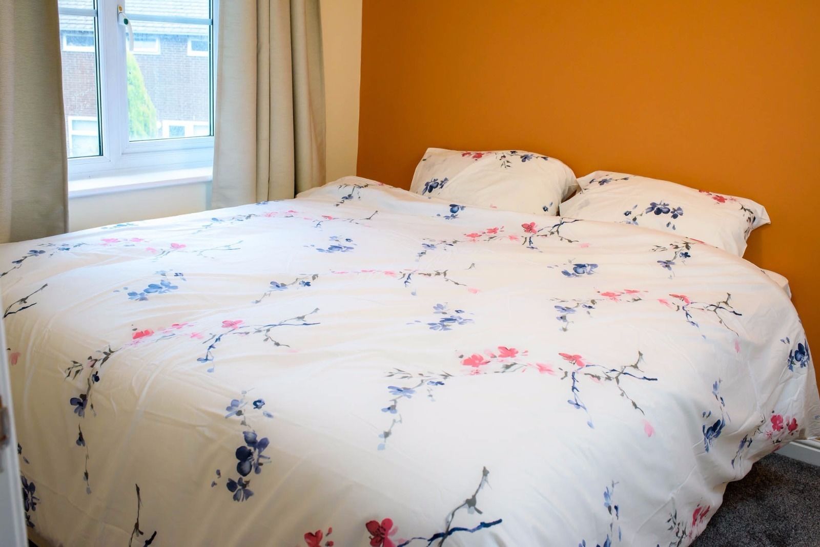 A bed with a floral bedspread in a room with an orange wall and a window with curtains.