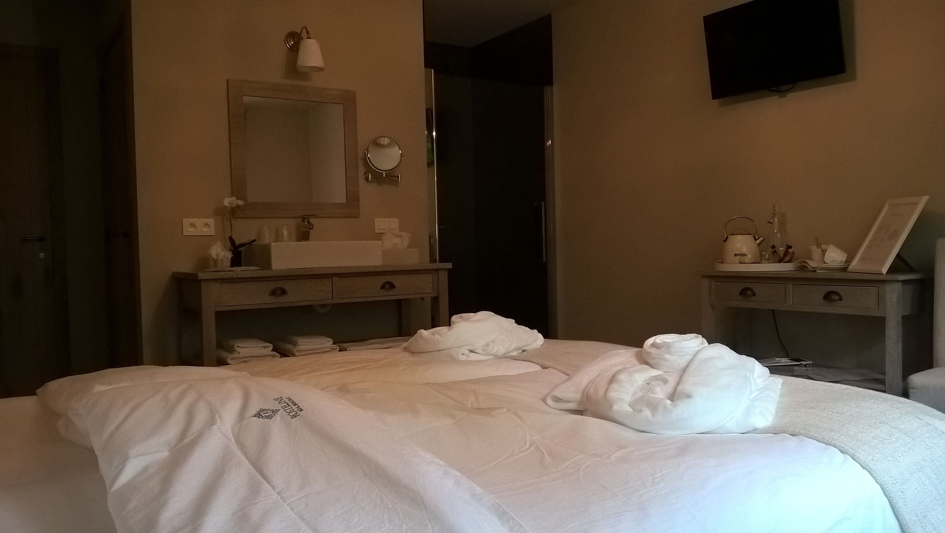 Et hotelværelse med en seng, et natbord med lampe, et spejl og et skrivebord med elkedel og billedramme.
