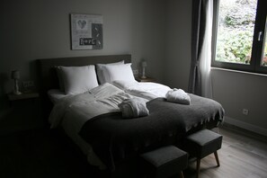 A neatly made bed with white linens and a gray blanket, a bedside table with a lamp, and a window with curtains. - B&B Rosteline (Hamont-Achel)