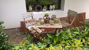 Un área de estar al aire libre de estilo rústico con una mesa y sillas de madera, rodeada de follaje y objetos decorativos.