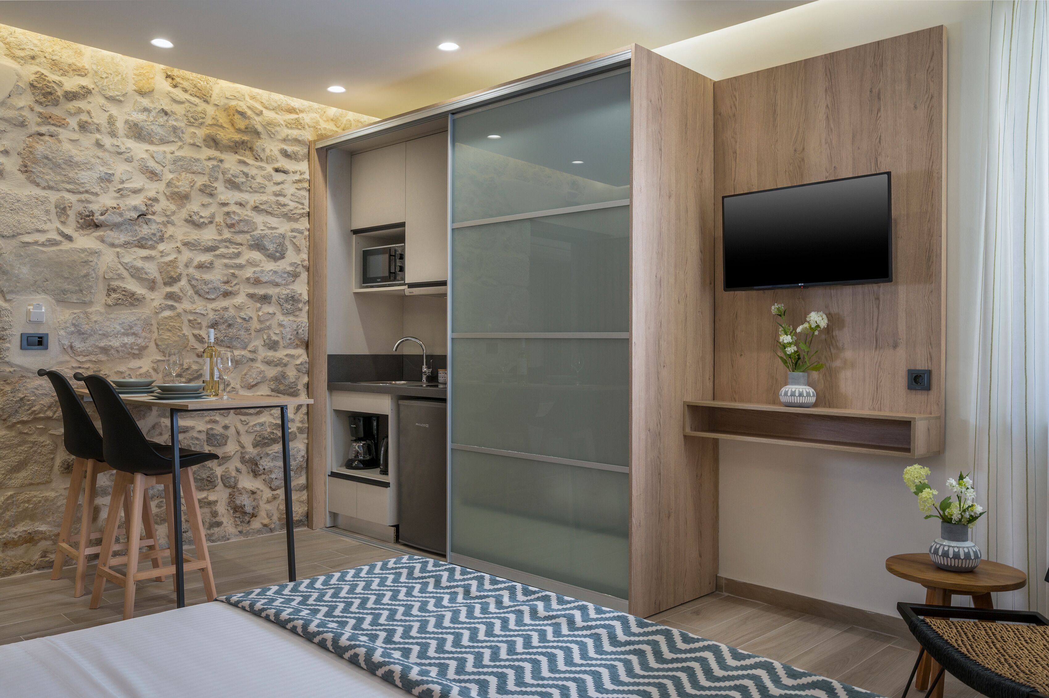 A compact room with a kitchenette, a dining area with chairs, a bed, and a TV mounted on the wall.