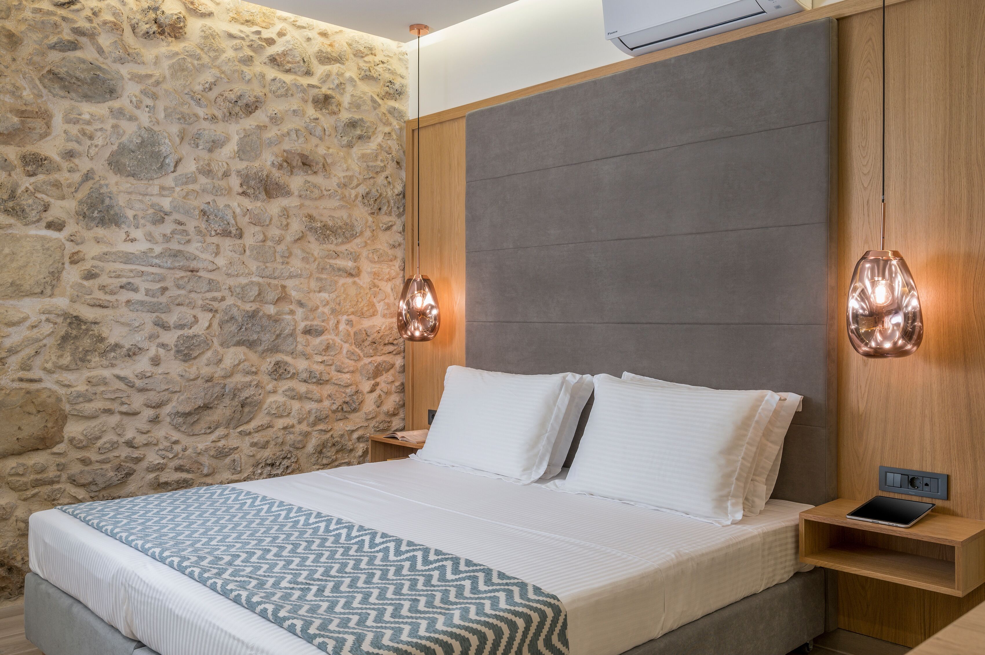 Quarto com parede de pedra, cama com roupa de cama branca e duas luminárias de cabeceira.
