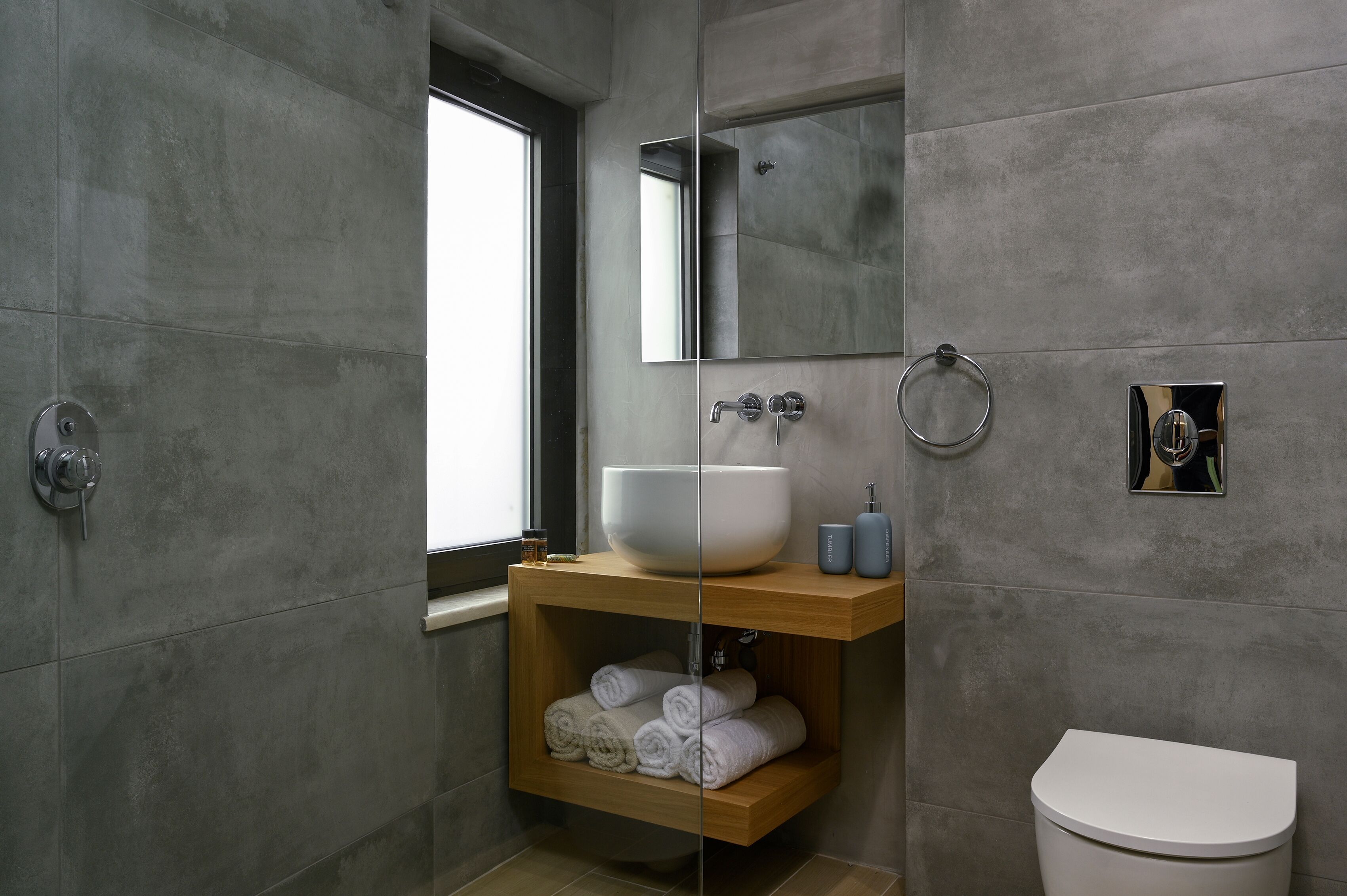Un bagno moderno con un ampio lavandino bianco, uno scaffale in legno con asciugamani, un WC e uno specchio.