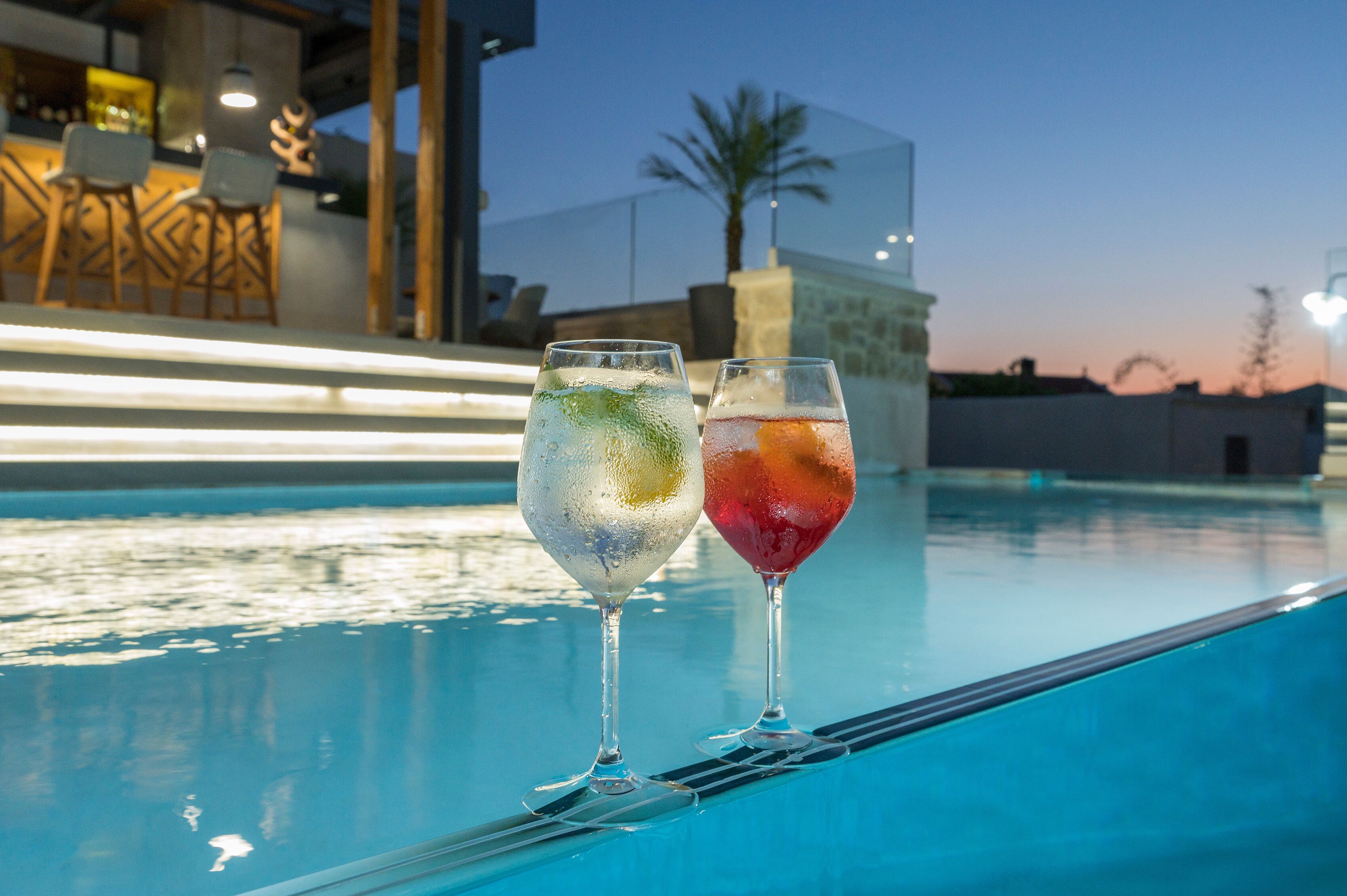 Two glasses of beverages on a poolside table with a bar and chairs in the background.