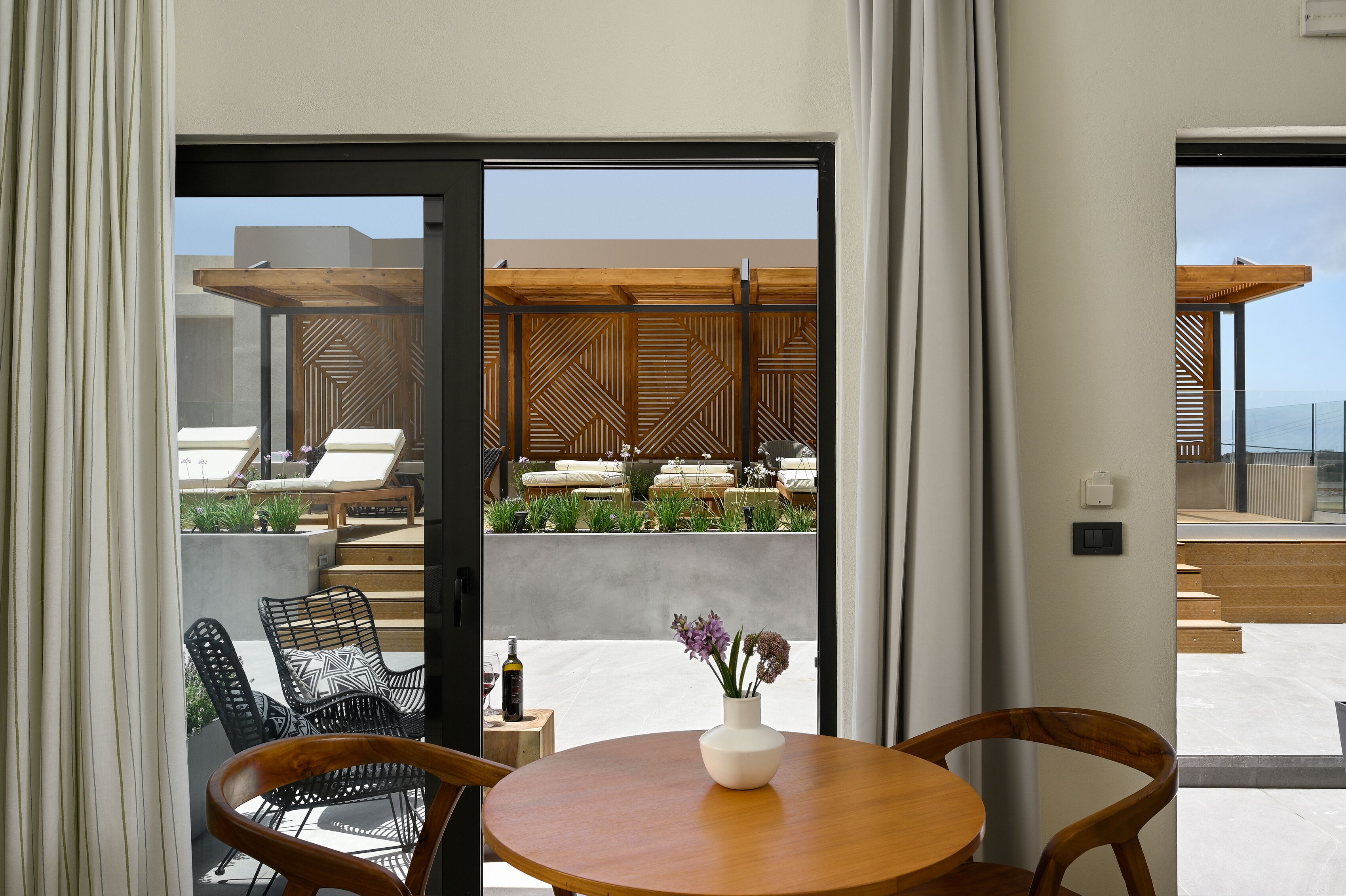 A modern room with a wooden table, chairs, and a view of an outdoor seating area with a geometric wooden pergola and plants.