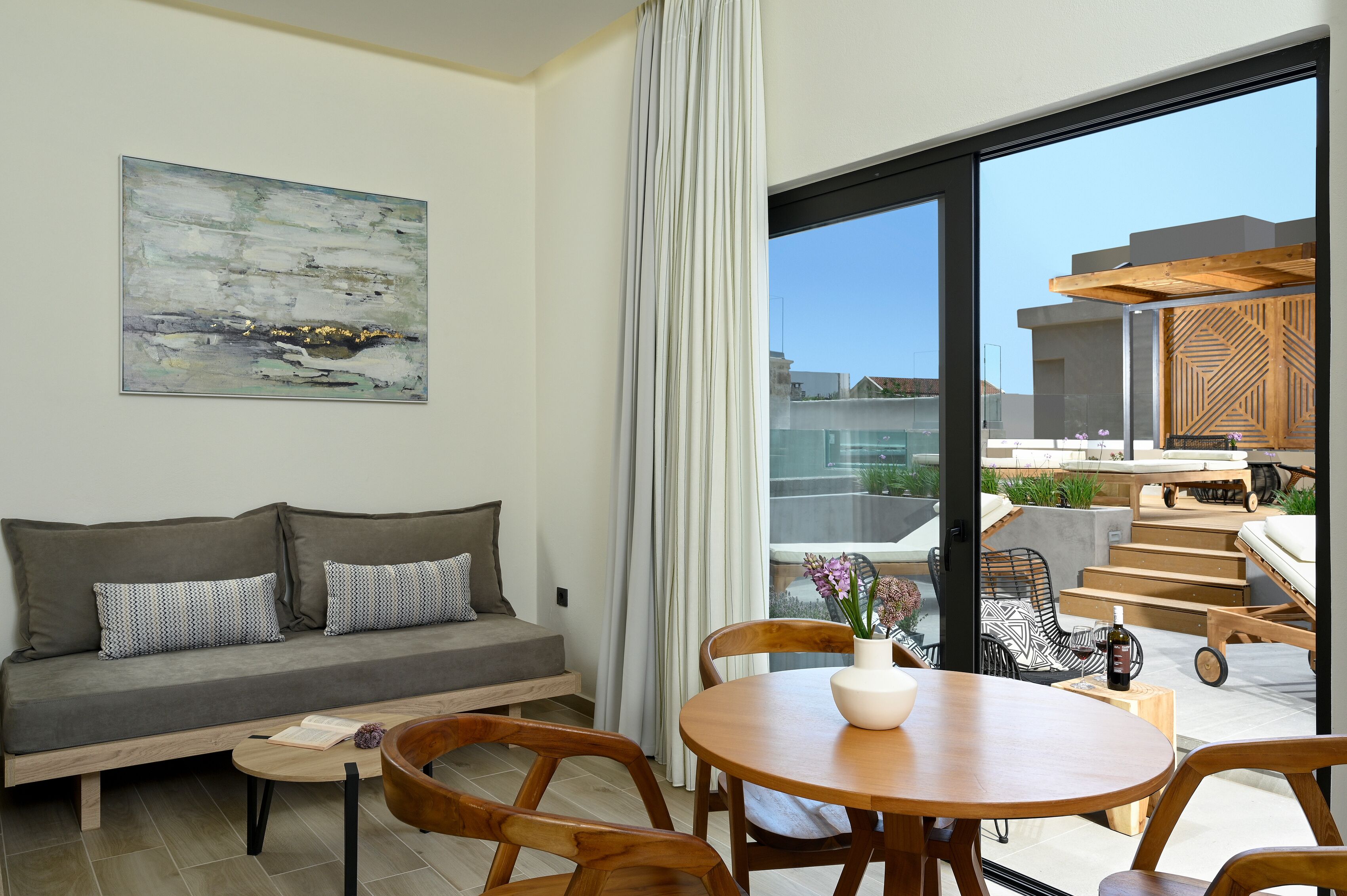 A modern living room with a grey sofa, wooden chairs, a round wooden table, and a large window with a view of the city.
