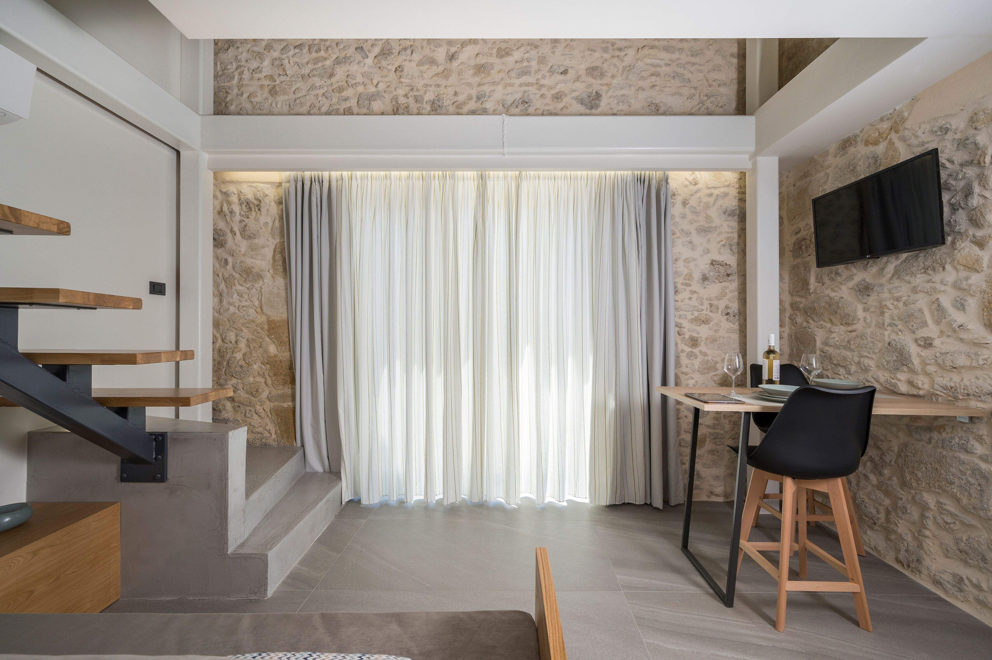 Un interior moderno con una escalera, un comedor con mesa y sillas, y un televisor montado en una pared de piedra.