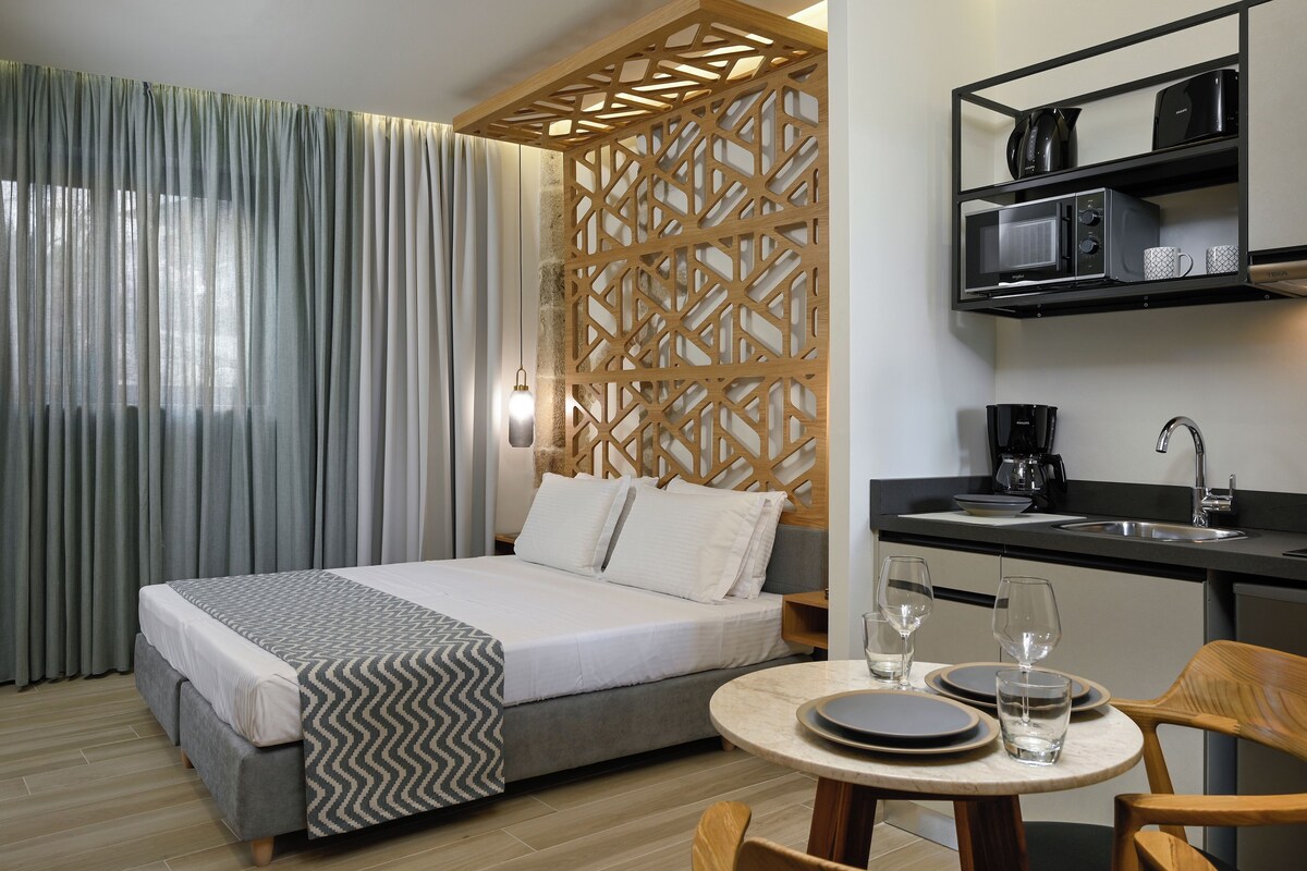 A hotel room with a bed, a dining table with chairs, a kitchen area with a microwave, and a decorative wooden partition.