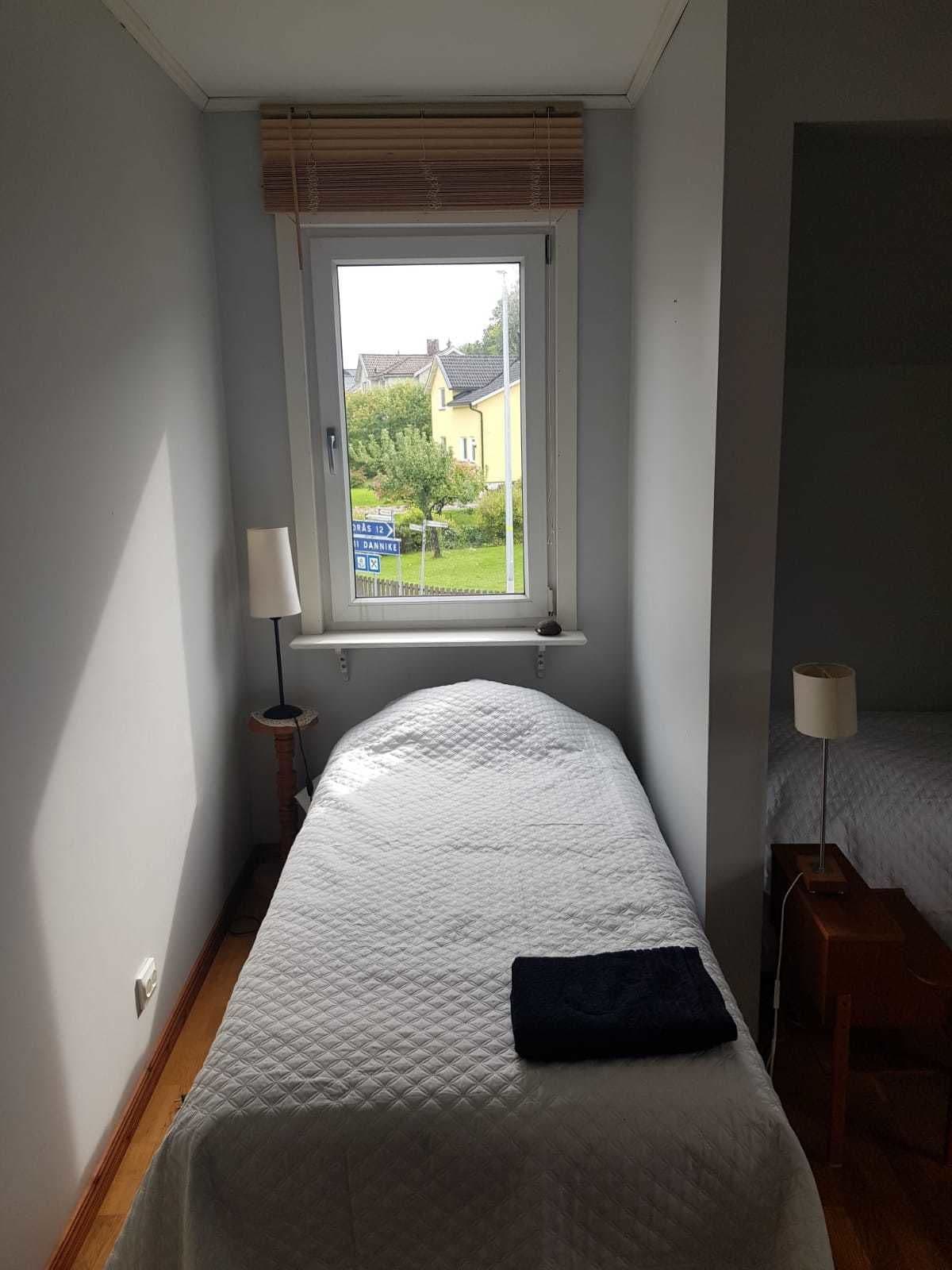 Quarto com cama, janela com persianas, abajur e vista para uma casa e vegetação lá fora.