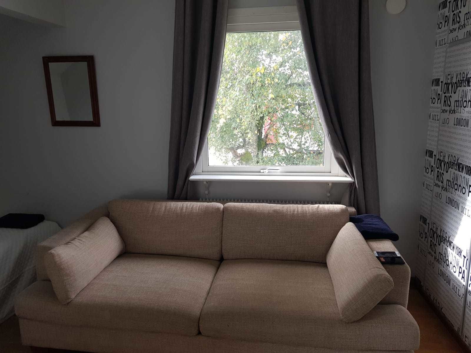 A living room with a beige sofa, a window with curtains, and a wall with printed text.