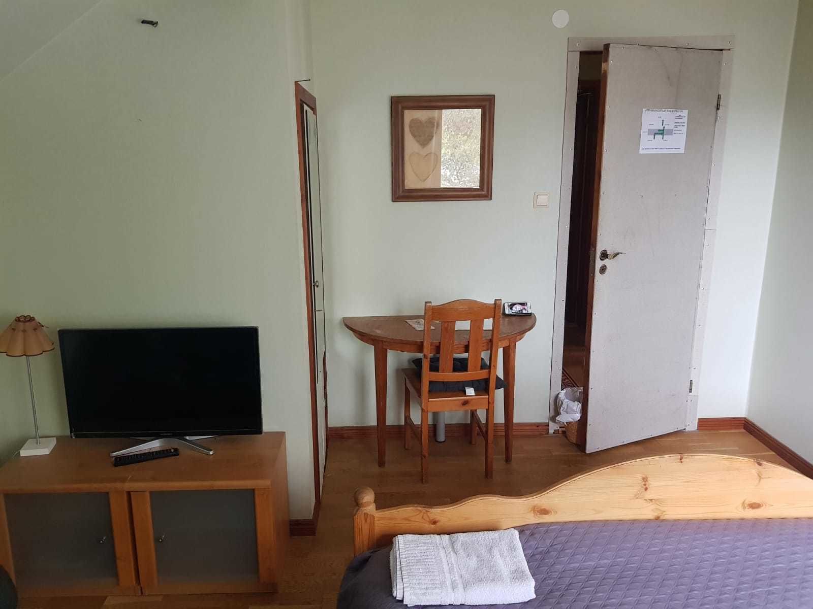 Ein kleiner Raum mit einem Bett, einem Fernseher auf einem Holzgestell, einem Tisch mit einem Stuhl und einer Tür mit einem Schild.