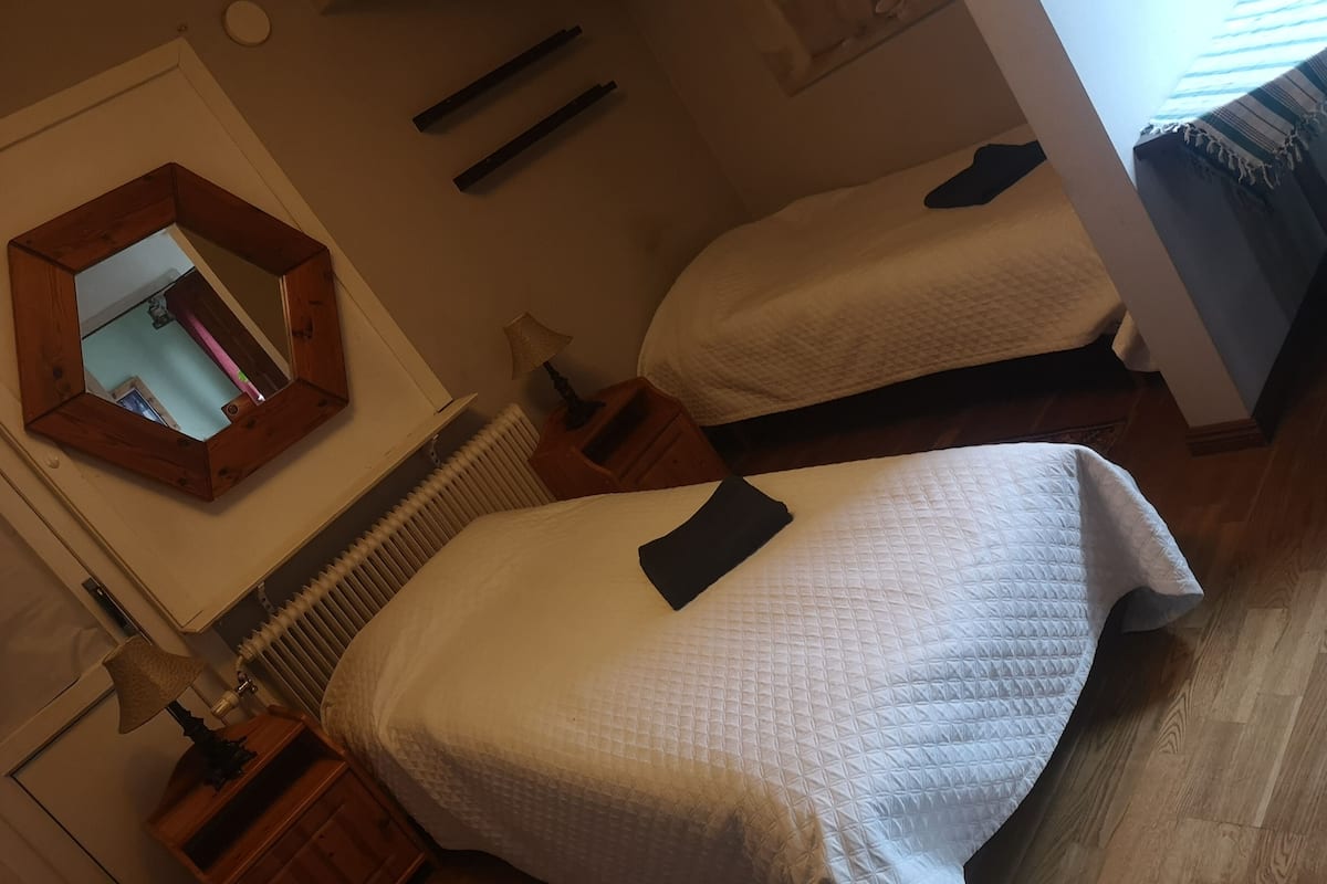 A bedroom with two beds, a wooden headboard, a mirror, a radiator, and a small table with a lamp.