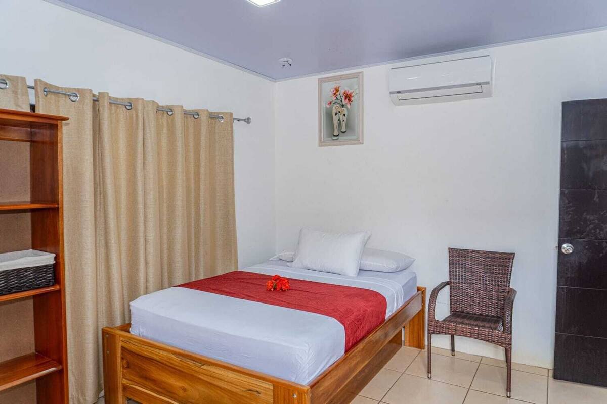 A bedroom with a wooden bed, a red blanket, a wicker chair, a shelf with baskets, and a wall-mounted air conditioner.