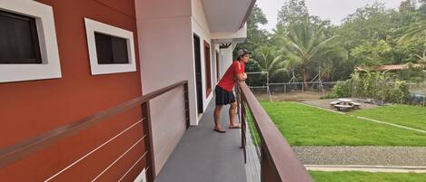 A balcony with a red building, a person leaning on the railing, and a green lawn with trees in the background.