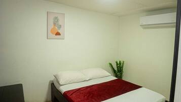 A bedroom with a bed, a red blanket, a white pillow, a small desk, a potted plant, and an air conditioning unit.