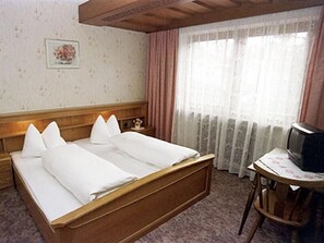 Une chambre d’hôtel avec un lit en bois, un petit bureau avec une télévision et une fenêtre avec des rideaux.